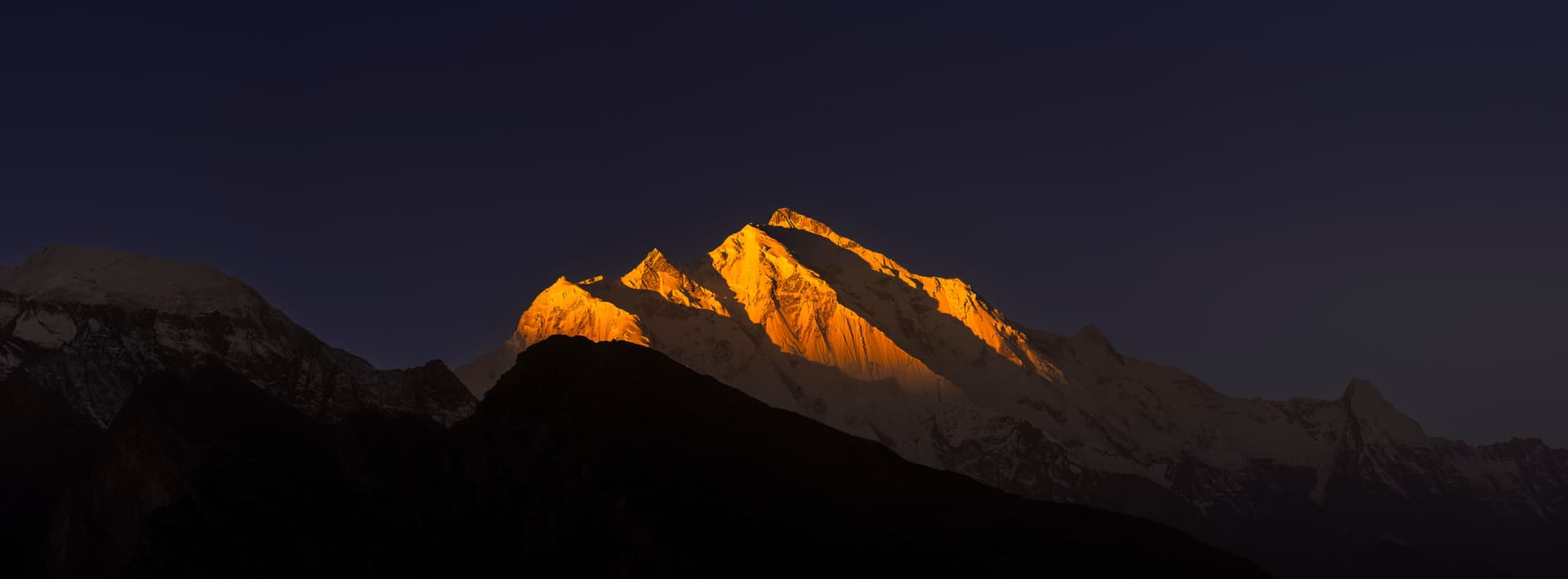 Dramatic photograph of a snow-capped mountain peak bathed in golden dawn light, rising above dark, shadowed ridges beneath a deep indigo sky.